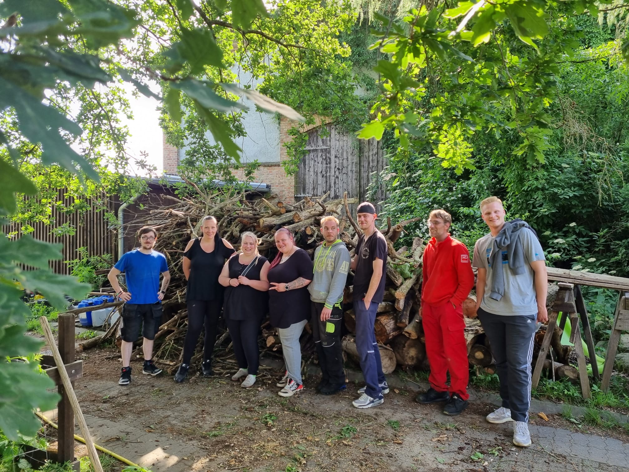 Eine Gruppe von sieben erwachsenen Helfern steht lächelnd vor einem großen Stapel geschnittenem Brennholz/Ästen.